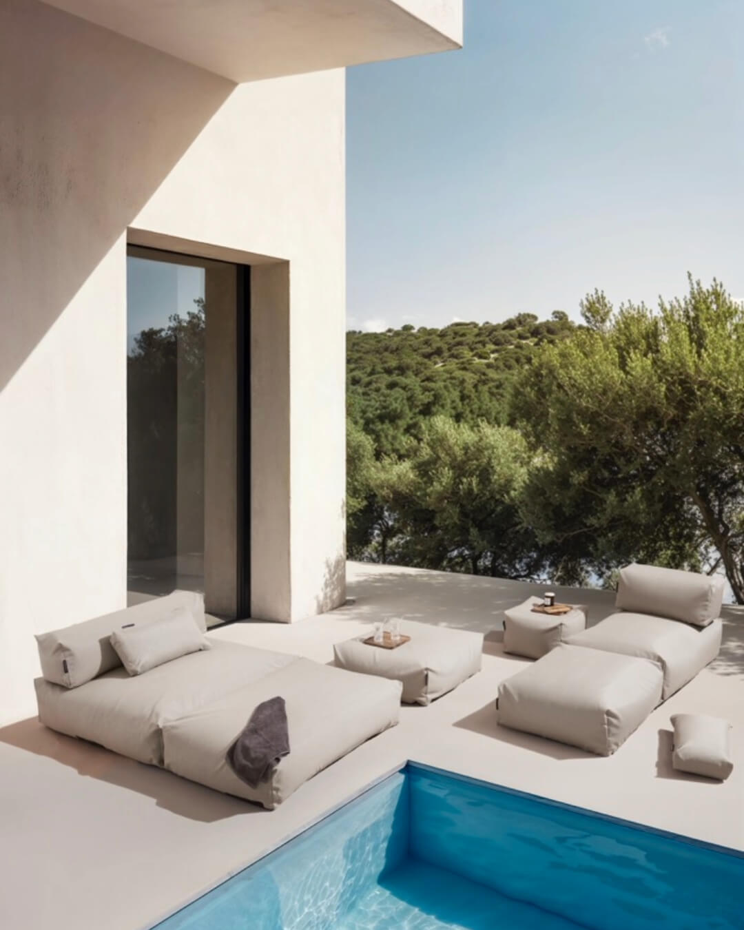 An einem Pool stehen zwei Poolsofas (ein Double, ein Single in Beige) zusammen und die Poufs in Large und Small sind als Tisch und weitere Sitzgelegenheit dazugestellt. $ Poolsofa Double & Single | Beige