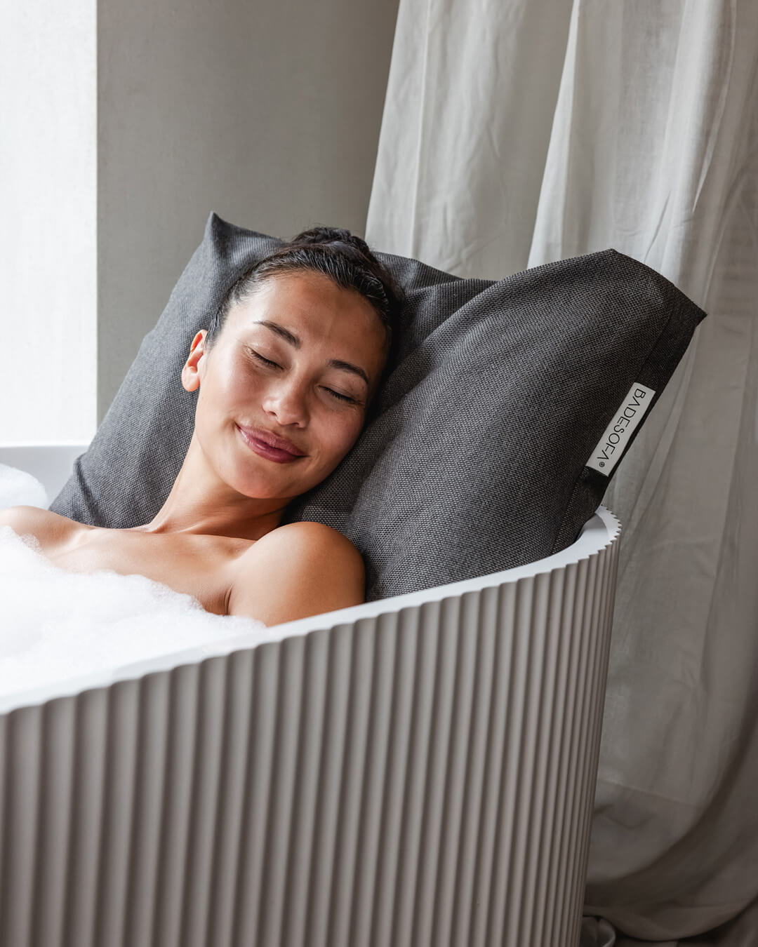 Mocha Badesofa Rückenkissen (Size Large) in einer freistehenden Badewanne, mit lächelnder Person, die sich entspannt anlehnt.