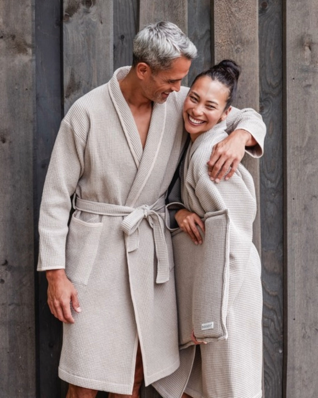Mann und Frau stehen im Bademantel (Farbe Anthrazit) vor einer Wand und lachen entspannt. 