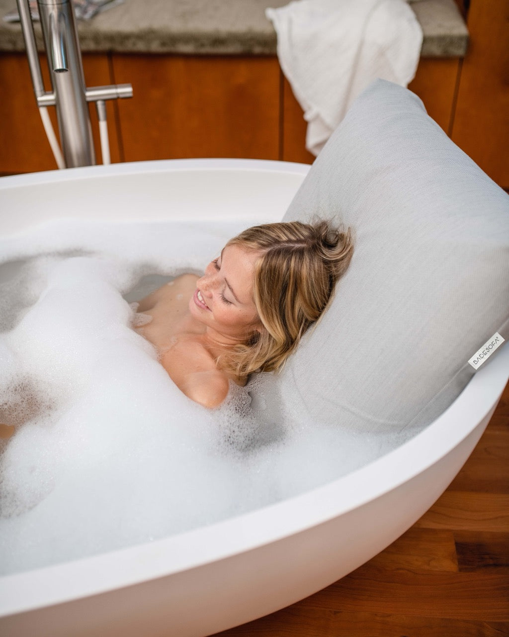Hellgraues Badesofa Rückenkissen (Size Large) in einer freistehenden Badewanne, mit lächelnder Person, die sich entspannt anlehnt.