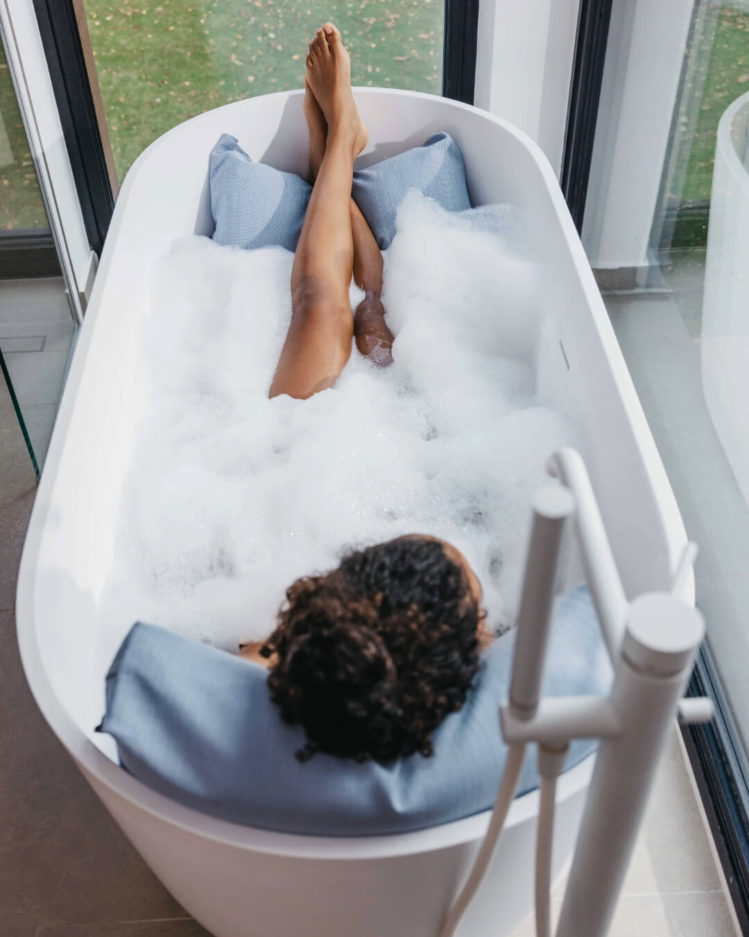 Frau badet mit Rückenkissen (Large, Blau) in luxuriöser freistehender Badewanne. Zusätzlich ruhen ihre Füße entspannt auf einem Badesofa Fusskissen (Farbe Blau). 