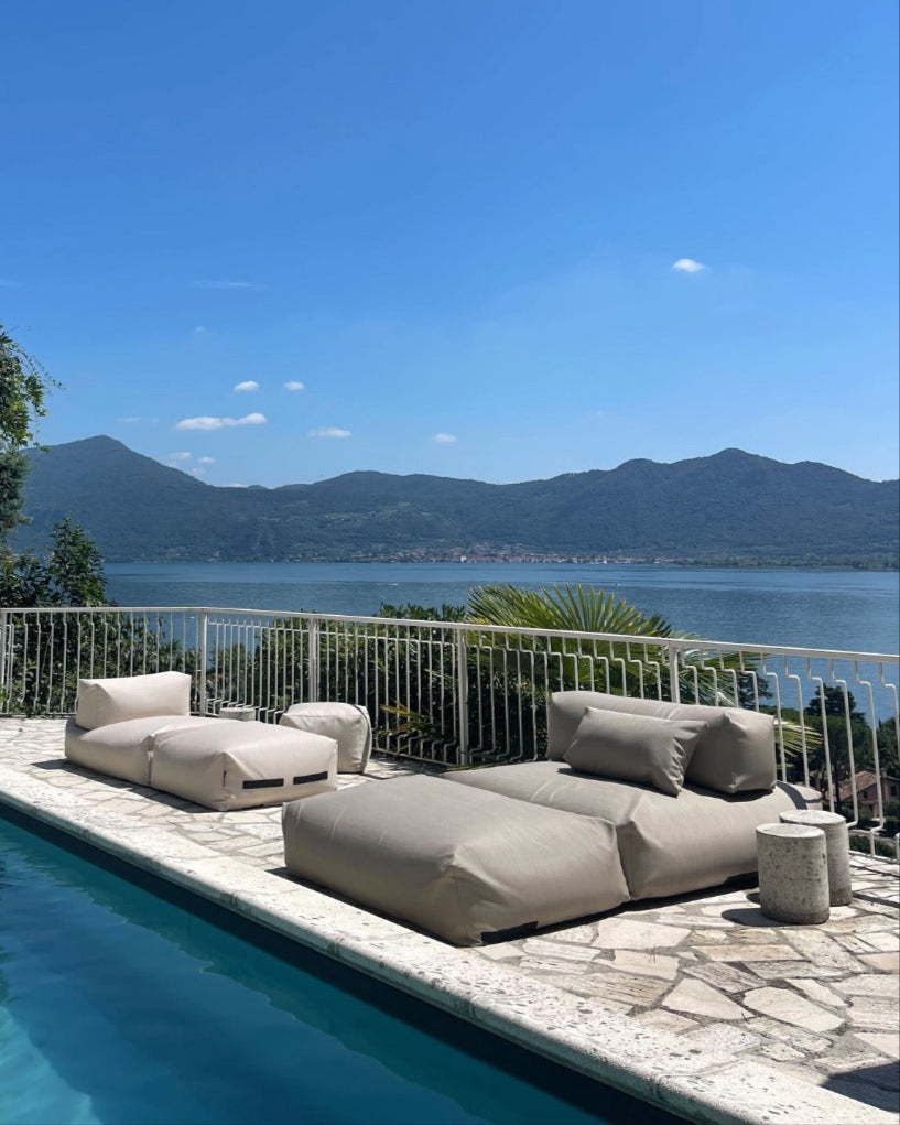 An einem Pool stehen zwei Poolsofas (ein Double in Beige, ein Single in Perlmutt) zusammen und ein Pouf in Small ist als Tisch dazugestellt. $ Poolsofa Double & Single | Perlmutt & Beige