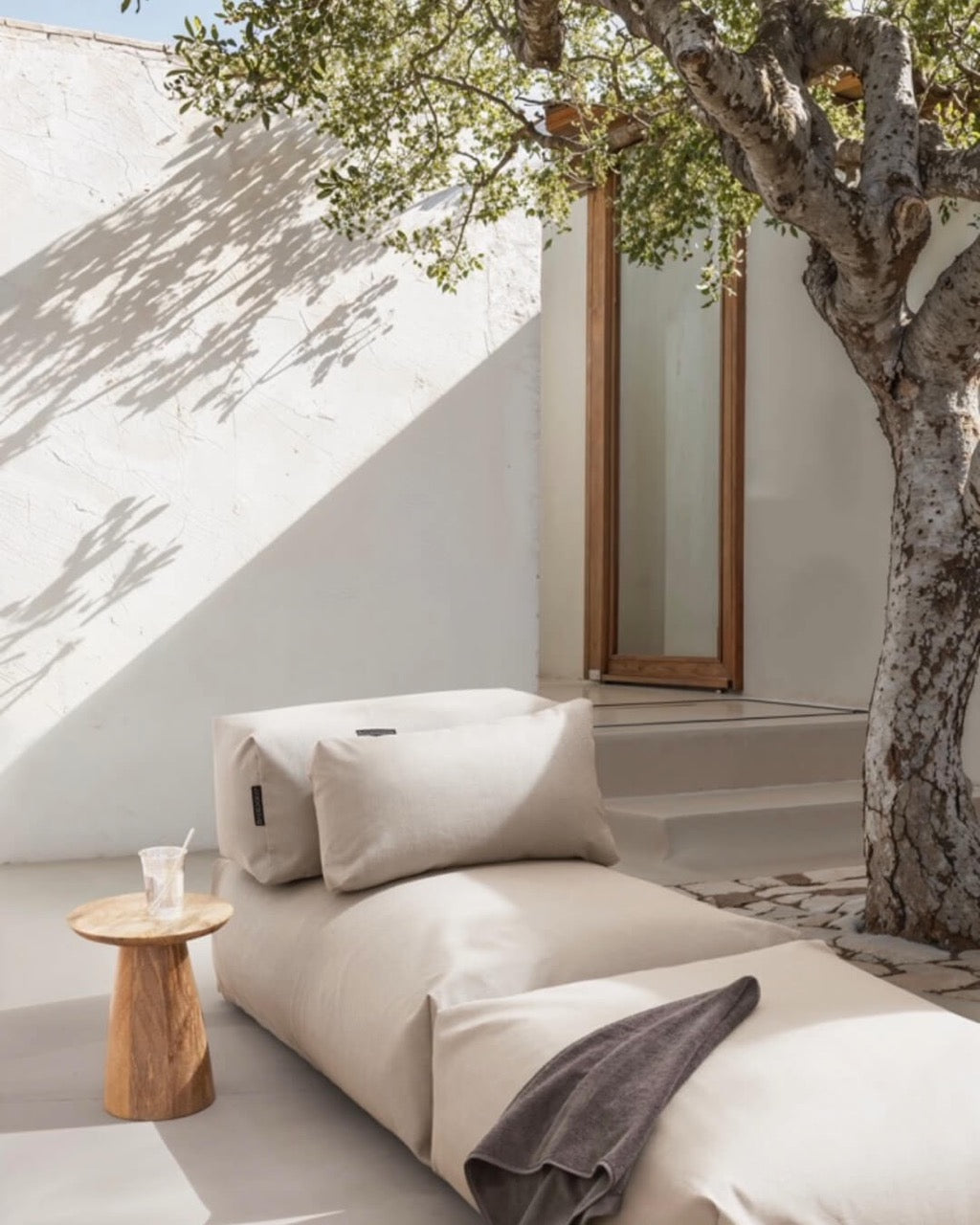 Unter einem Baum steht ein Poolsofa Singel in Beige, daneben ein kleiner Holztisch. Das Rückenkissen und Relaxkissen sind auf dem Poolsofa drapiert. $ Poolsofa Single | Beige