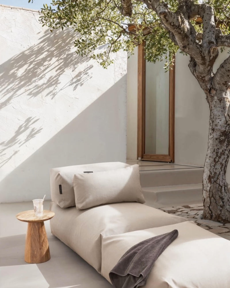 Unter einem Baum steht ein Poolsofa Singel in Beige, daneben ein kleiner Holztisch. Das Rückenkissen und Relaxkissen sind auf dem Poolsofa drapiert. 