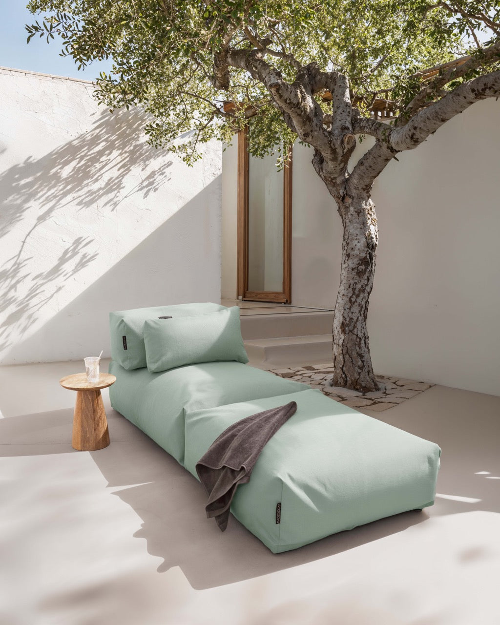 Unter einem Baum steht ein Poolsofa Singel in Grün, daneben ein kleiner Holztisch. Das Rückenkissen und Relaxkissen sind auf dem Poolsofa drapiert.