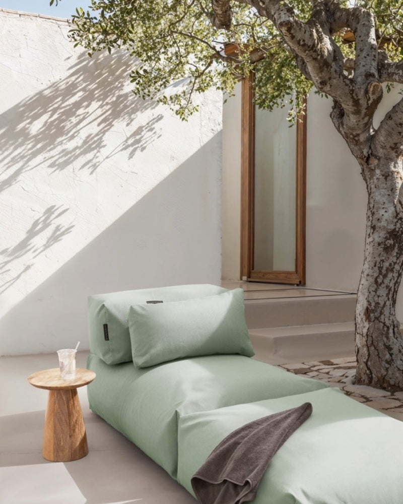 Unter einem Baum steht ein Poolsofa Singel in Grün, daneben ein kleiner Holztisch. Das Rückenkissen und Relaxkissen sind auf dem Poolsofa drapiert. 