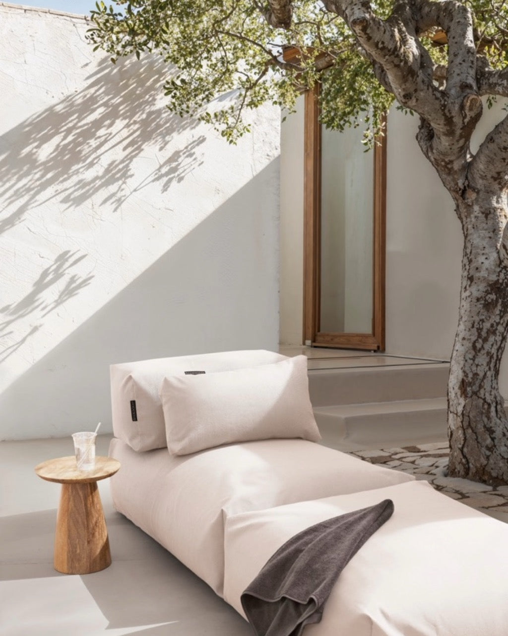 Unter einem Baum steht ein Poolsofa Singel in Perlmutt, daneben ein kleiner Holztisch. Das Rückenkissen und Relaxkissen sind auf dem Poolsofa drapiert. 