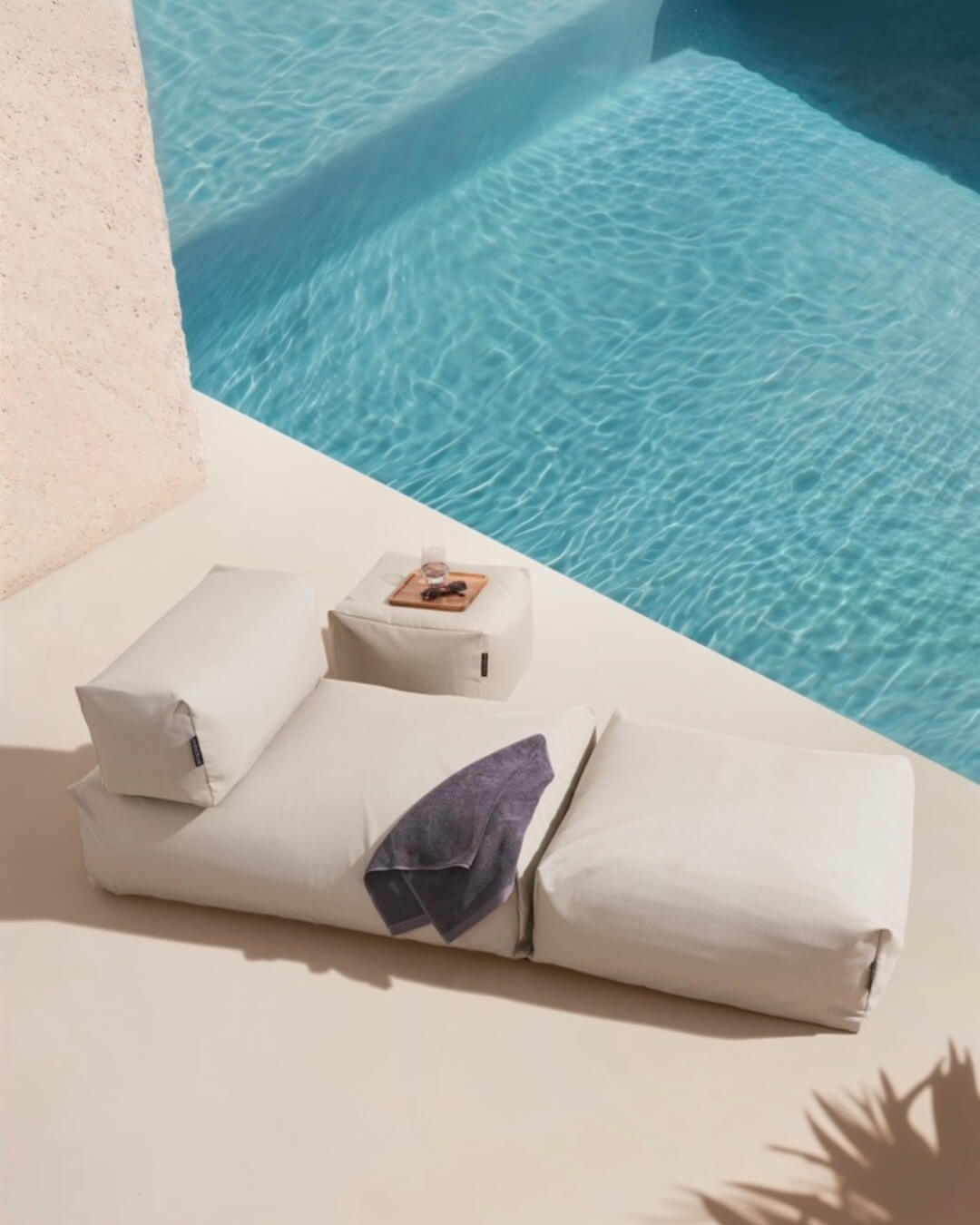 Ein Poolsofa single in PErlmutt steht neben einem Pool. Ein Pouf in small dient als Ablage und kleiner Tisch. $ Poolsofa Single + Pouf Small