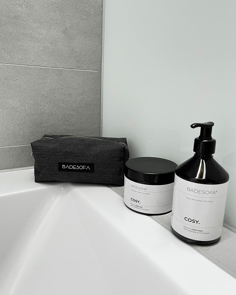 Kosmetiktasche steht neben einem Tiegel Badesalz und einer Flasche Badeschaum von Badesofa auf dem Badewannenrand.